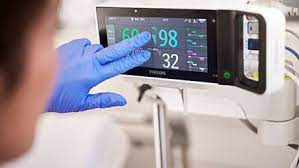 The image depicts a healthcare professional interacting with a Philips IntelliVue monitor, a Human-Machine Interface system used in hospitals to display real-time patient vital signs such as heart rate, oxygen saturation, and respiratory rate for continuous clinical monitoring.
